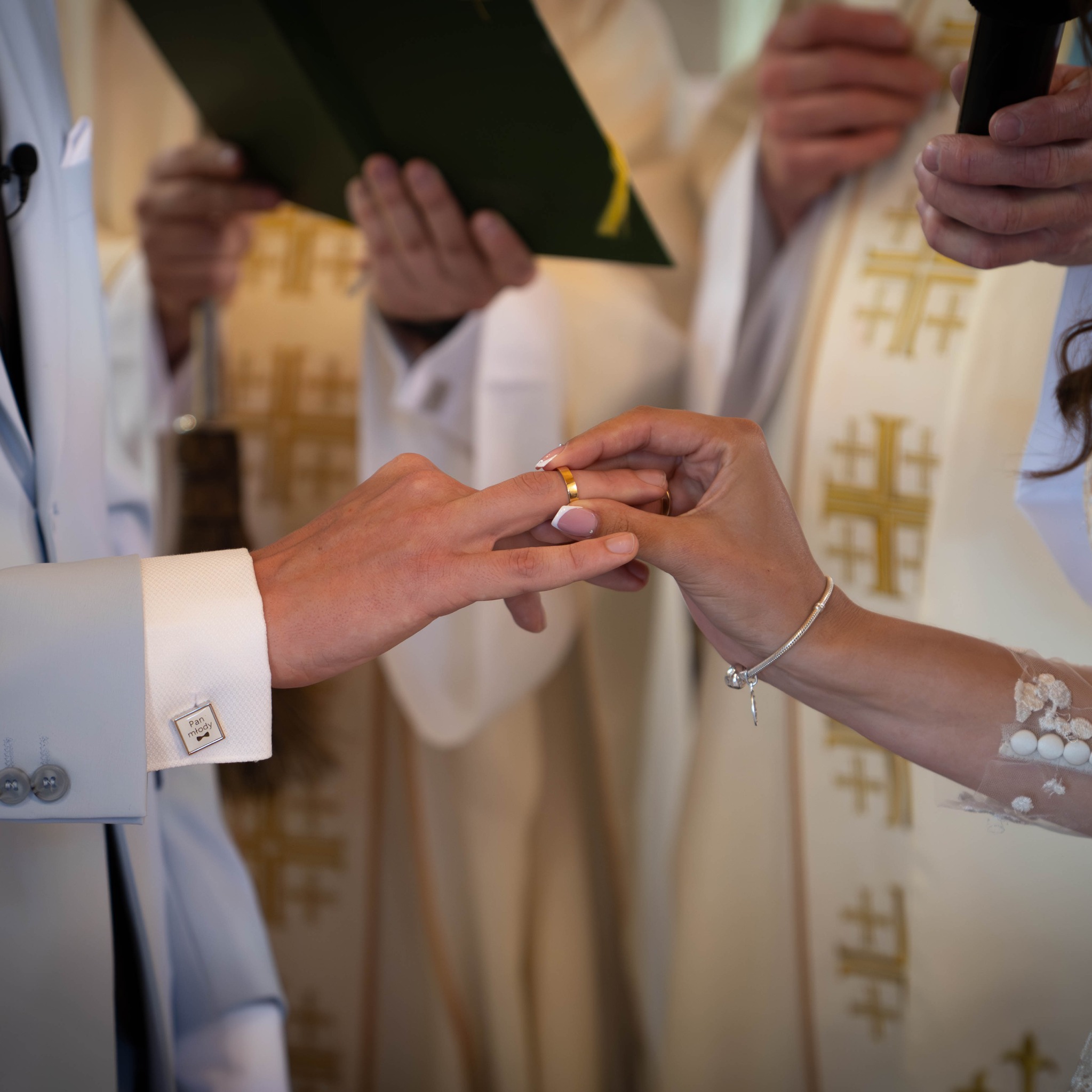 Moment zakładania obrączki podczas ceremonii ślubnej. Widoczne dłonie nowożeńców, fragment sukni panny młodej i mankiet koszuli pana młodego z napisem 'Pan młody'.