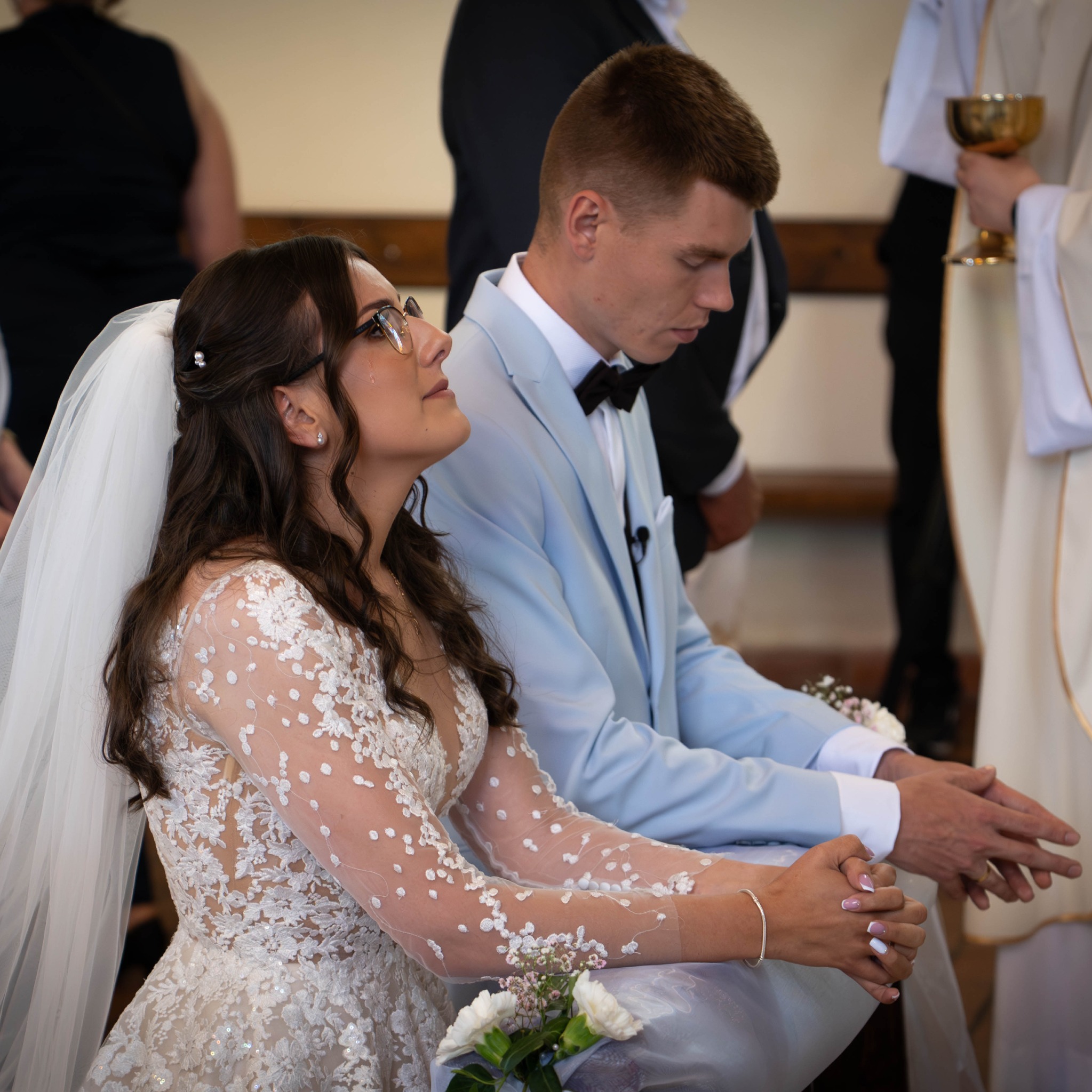 Uczestnicy ceremonii ślubnej: panna młoda w koronkowej sukni i pan młody w błękitnym garniturze klęczą obok księdza z kielichem. Ślub w kościele.
