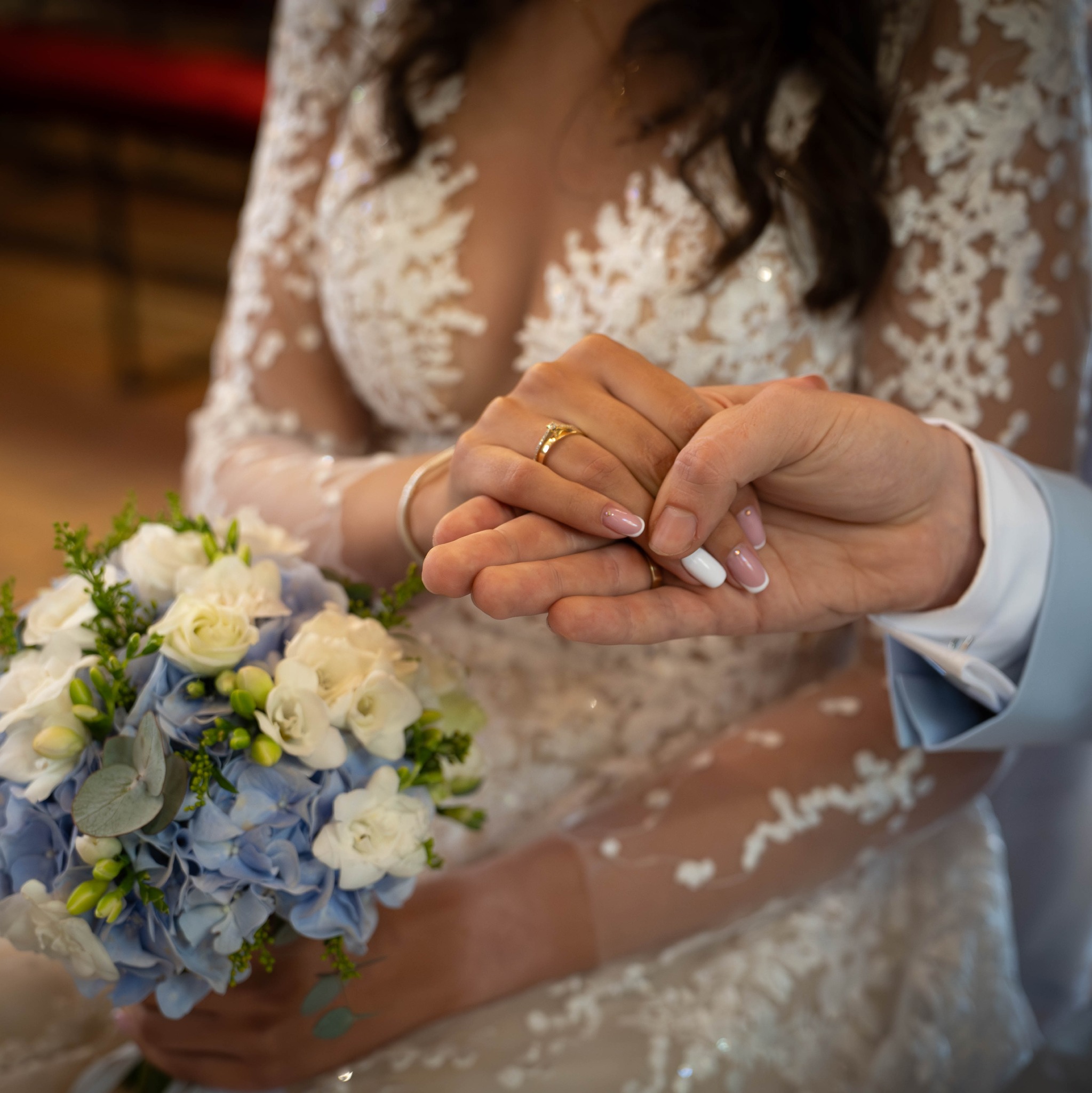 Zbliżenie dłoni nowożeńców z obrączkami na tle bukietu ślubnego i sukni z koronkowymi rękawami, delikatne światło podkreśla detale.