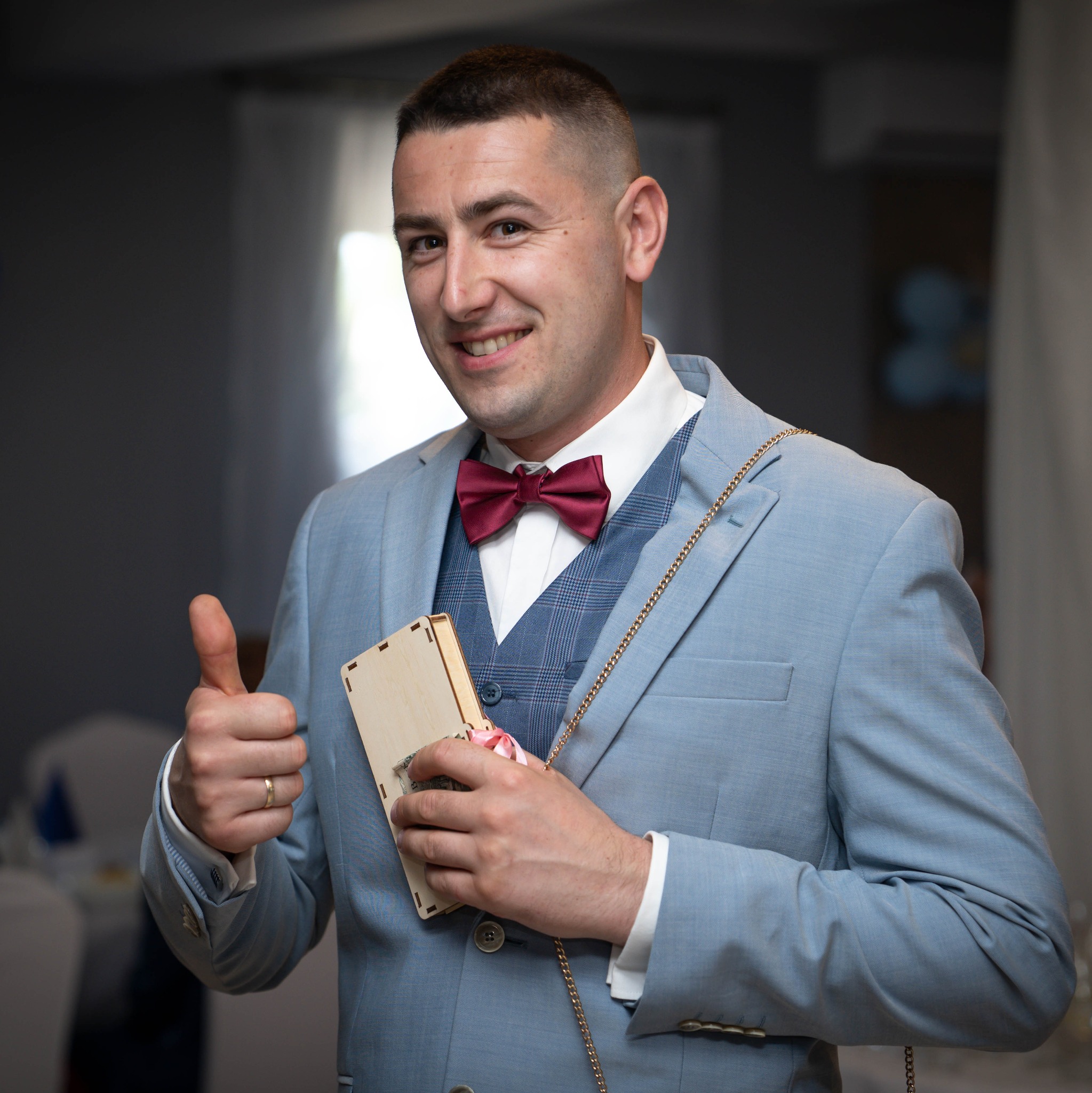 Uśmiechnięty mężczyzna w błękitnym garniturze i bordowej muszce, trzymający drewniane pudełko z pieniędzmi i pokazujący kciuk w górę na tle sali bankietowej.