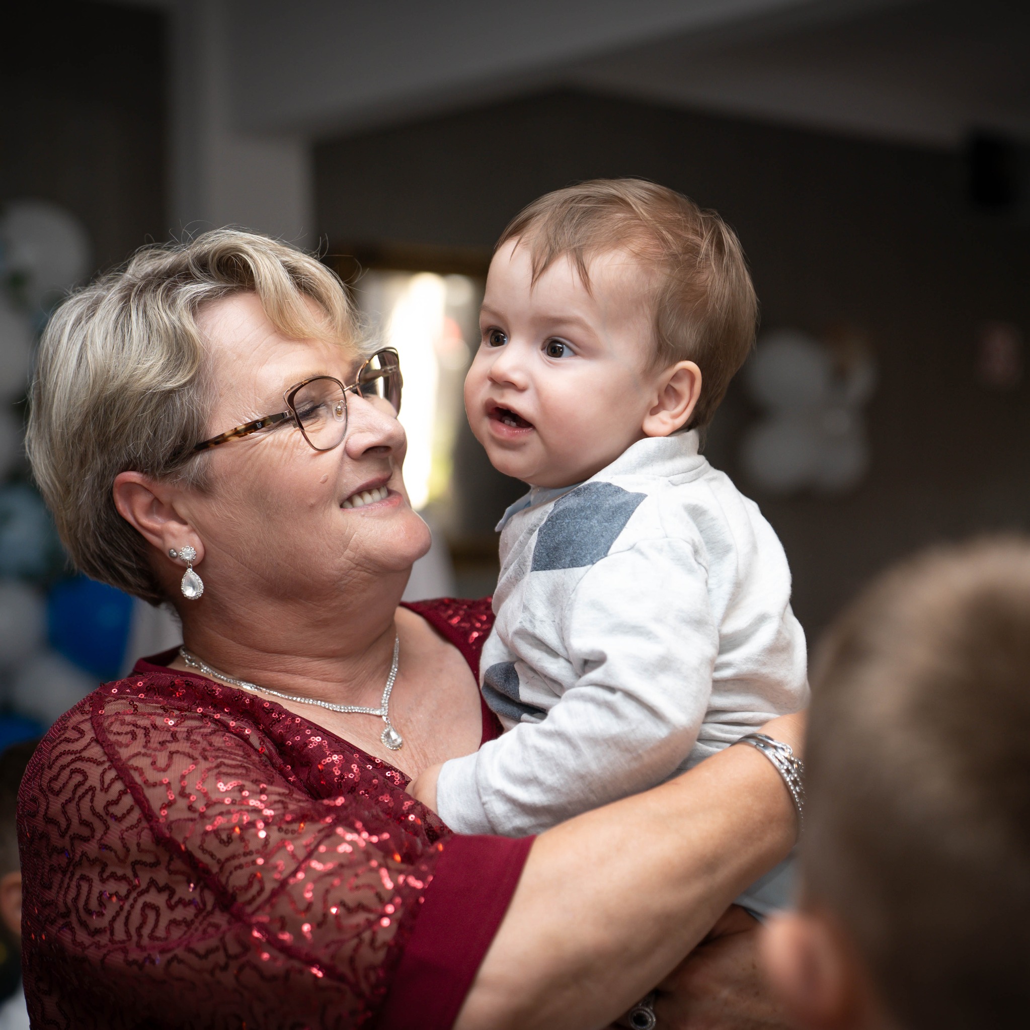 Uśmiechnięta kobieta w bordowej sukience trzyma na rękach małego chłopca, który patrzy w bok. W tle dekoracje.