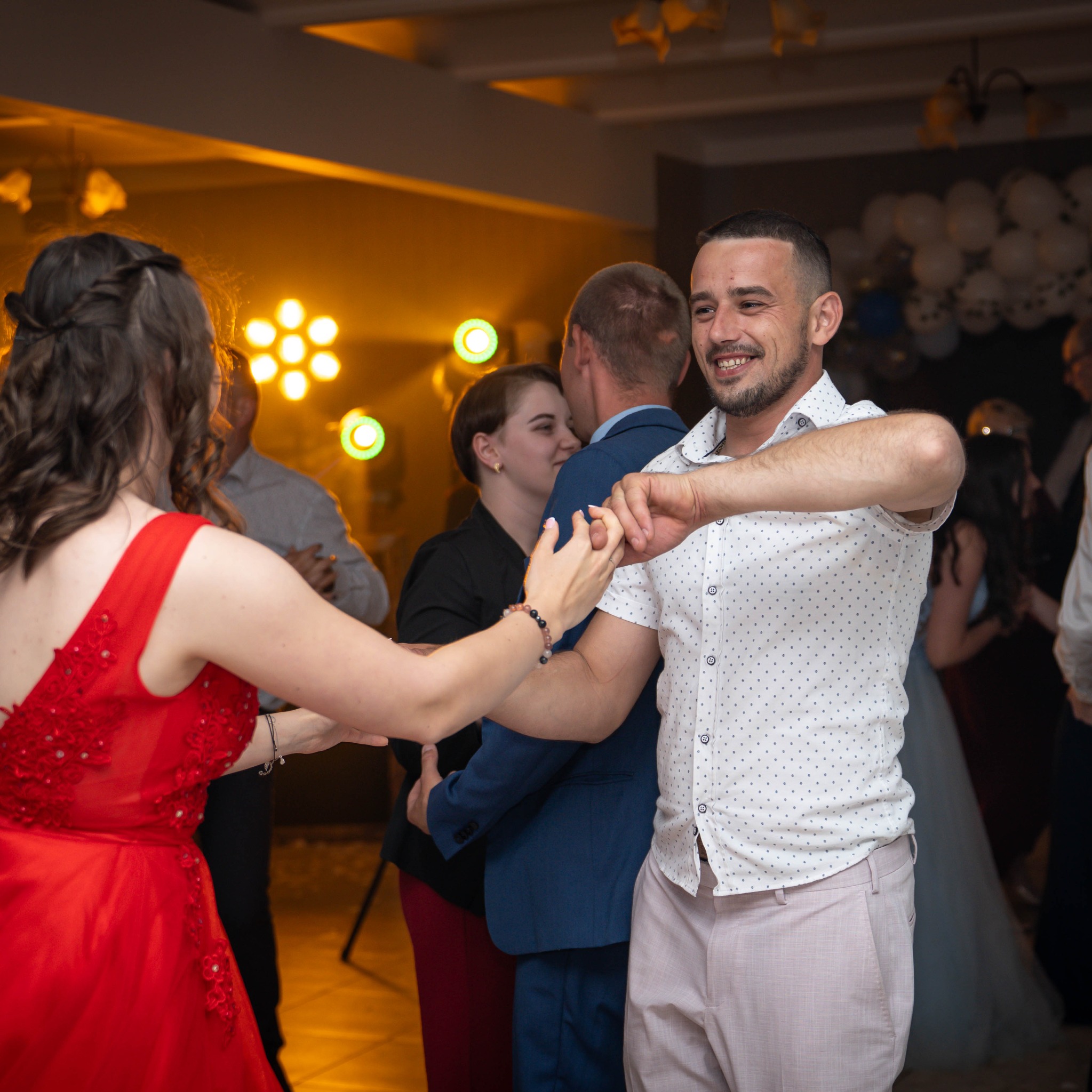Radosny taniec pary na tle dekoracji świetlnych i balonów. Mężczyzna w koszuli w kropki i różowych spodniach uśmiecha się, trzymając dłoń kobiety w czerwonej sukni.