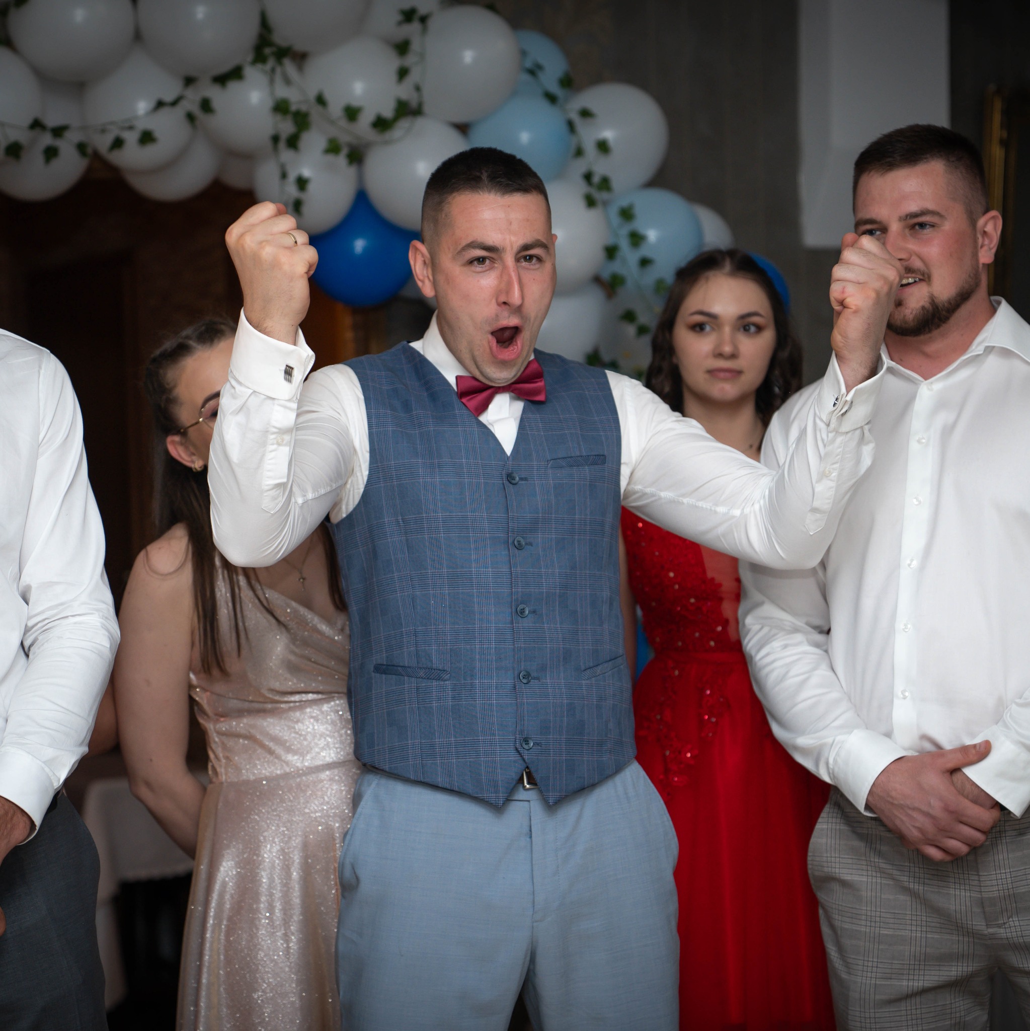 Rozradowany mężczyzna w muszce i kamizelce na tle dekoracji balonowych, świętujący w otoczeniu gości ubranych w eleganckie stroje.