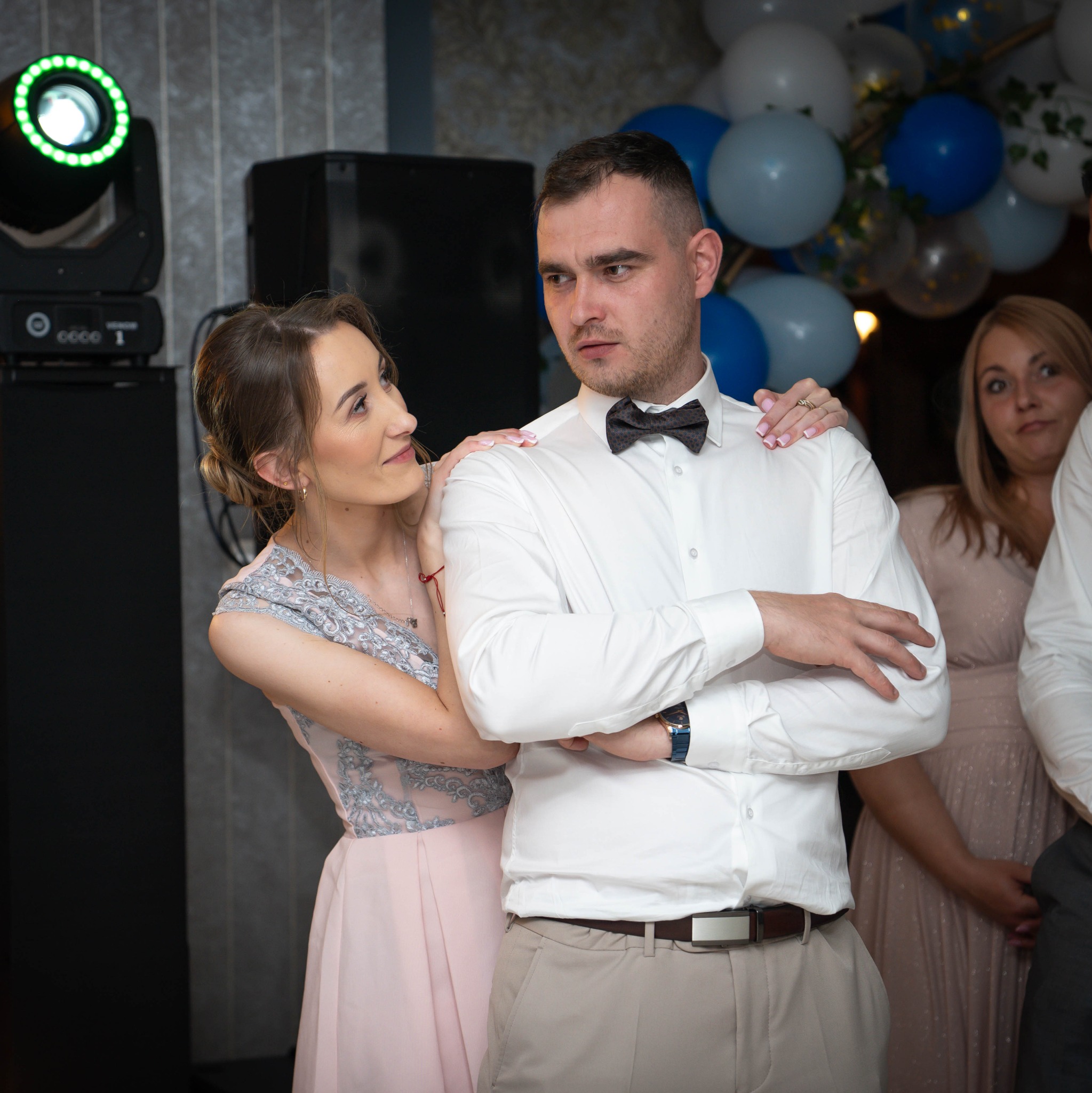 Uśmiechnięta kobieta w pudrowej sukni dotyka ramienia mężczyzny w białej koszuli i muszce na tle dekoracji balonowych i oświetlenia eventowego.