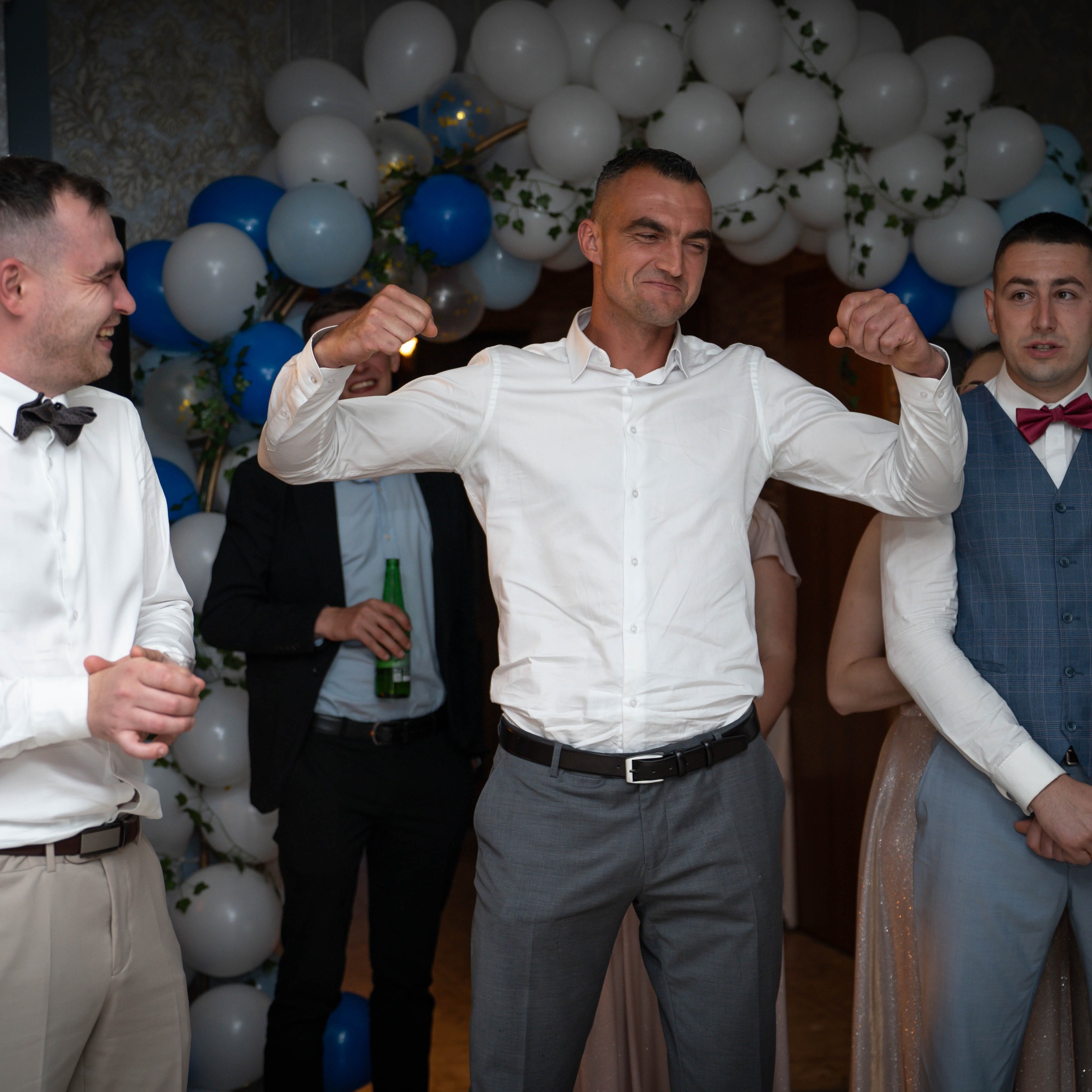 Radosna atmosfera na imprezie: mężczyzna w białej koszuli, z szerokim uśmiechem, pręży muskuły na tle dekoracji z białych i niebieskich balonów. Towarzyszą mu inni goście.