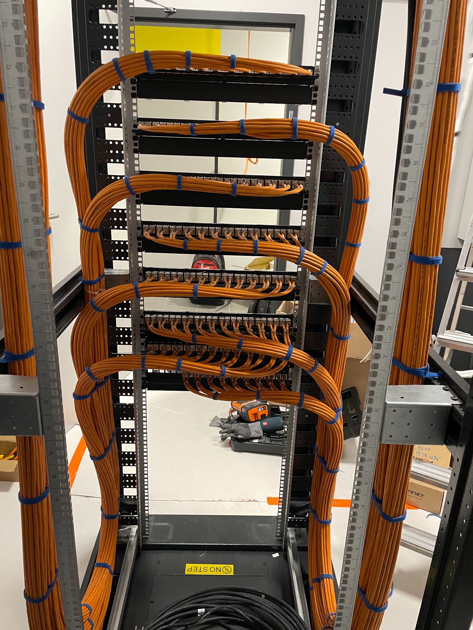 Rząd pomarańczowych kabli sieciowych, schludnie ułożonych w czarnej szafie rack, zabezpieczonych niebieskimi opaskami. Widoczny fragment podłogi i narzędzia w tle.