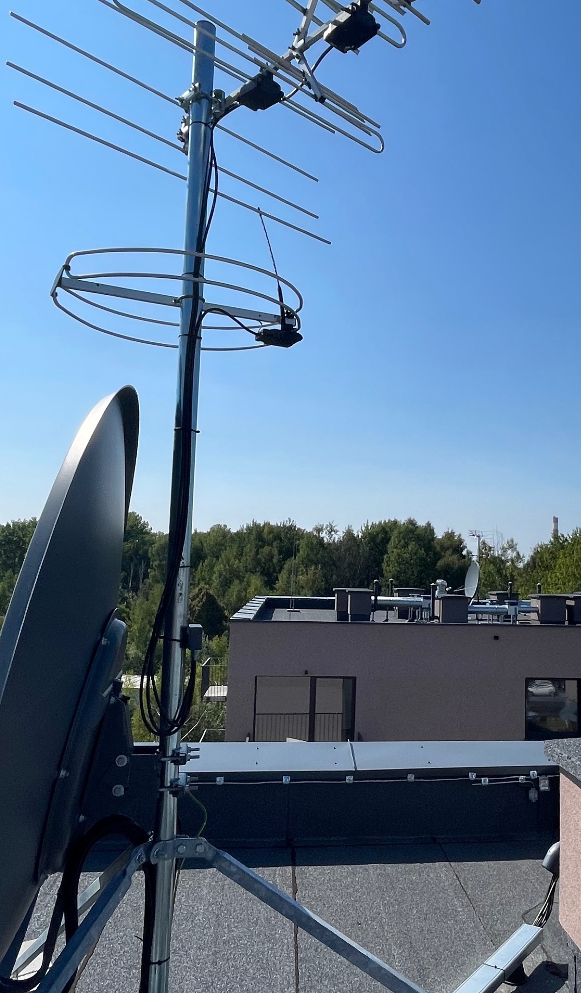 Antena telewizyjna i satelitarna na dachu budynku, kable starannie poprowadzone wzdłuż masztu, błękitne niebo w tle. Widok z dołu.