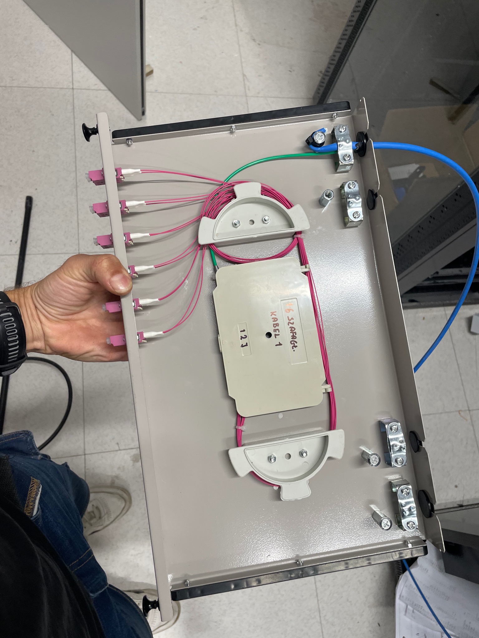 Panel światłowodowy z podłączonymi kablami i elementami mocującymi, trzymany przez osobę. Widoczne kable w kolorach różowym, zielonym i niebieskim.