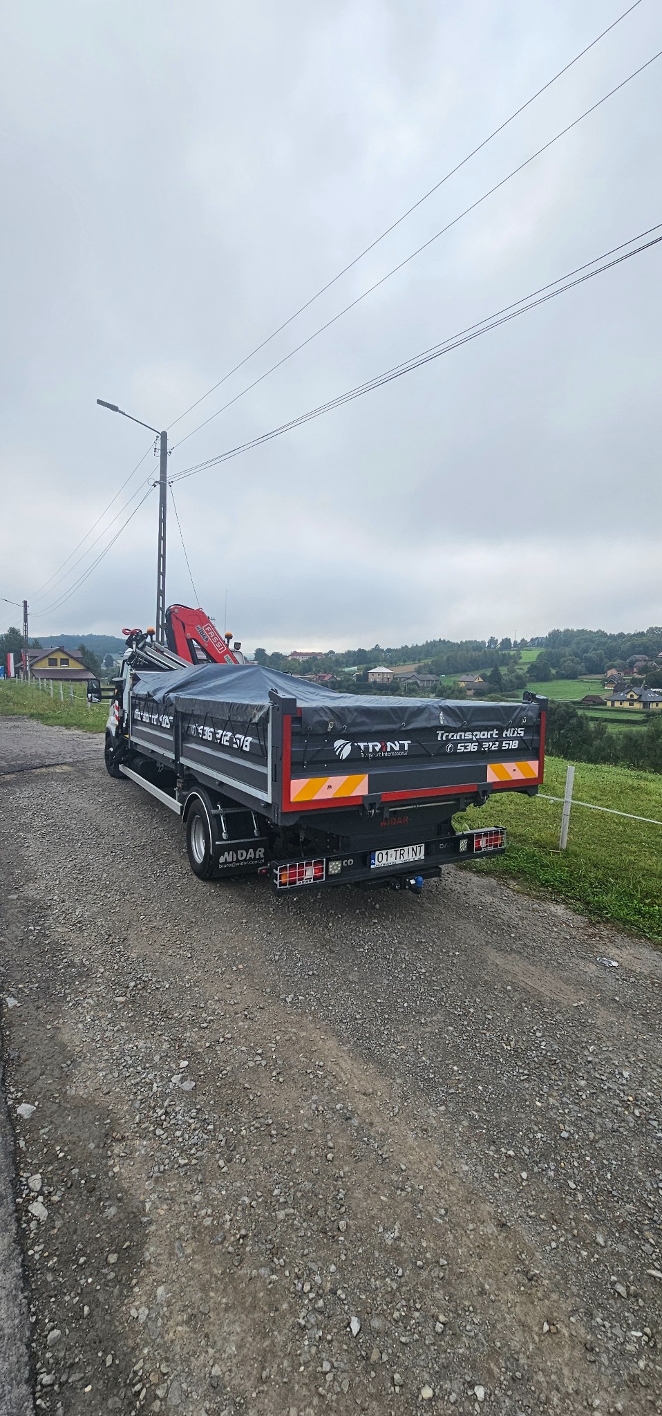 Ciężarówka z dźwigiem HDS marki Fassi, zaparkowana na żwirowej drodze w pochmurny dzień. Widoczny numer telefonu i logo firmy transportowej TRINT.
