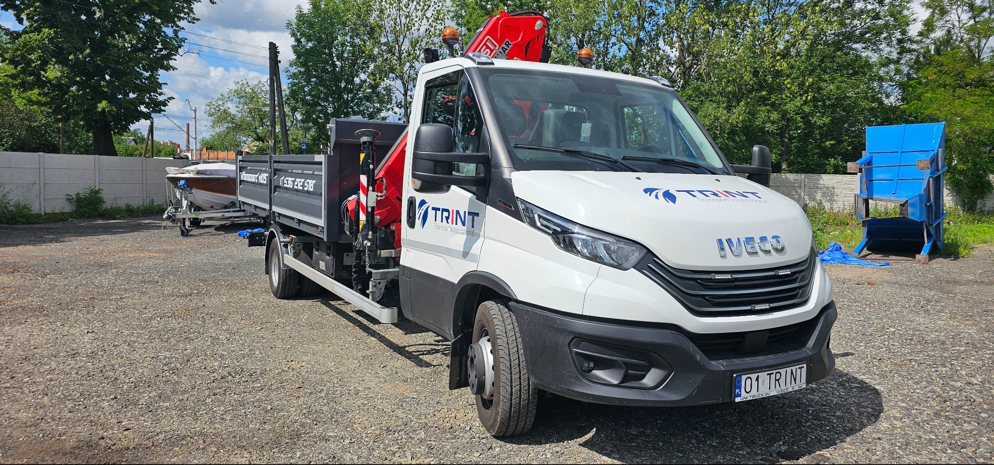 Biała laweta z dźwigiem marki Iveco, z logo firmy TRINT, zaparkowana na żwirowym placu. Na lawecie przewożona jest łódź na przyczepie.