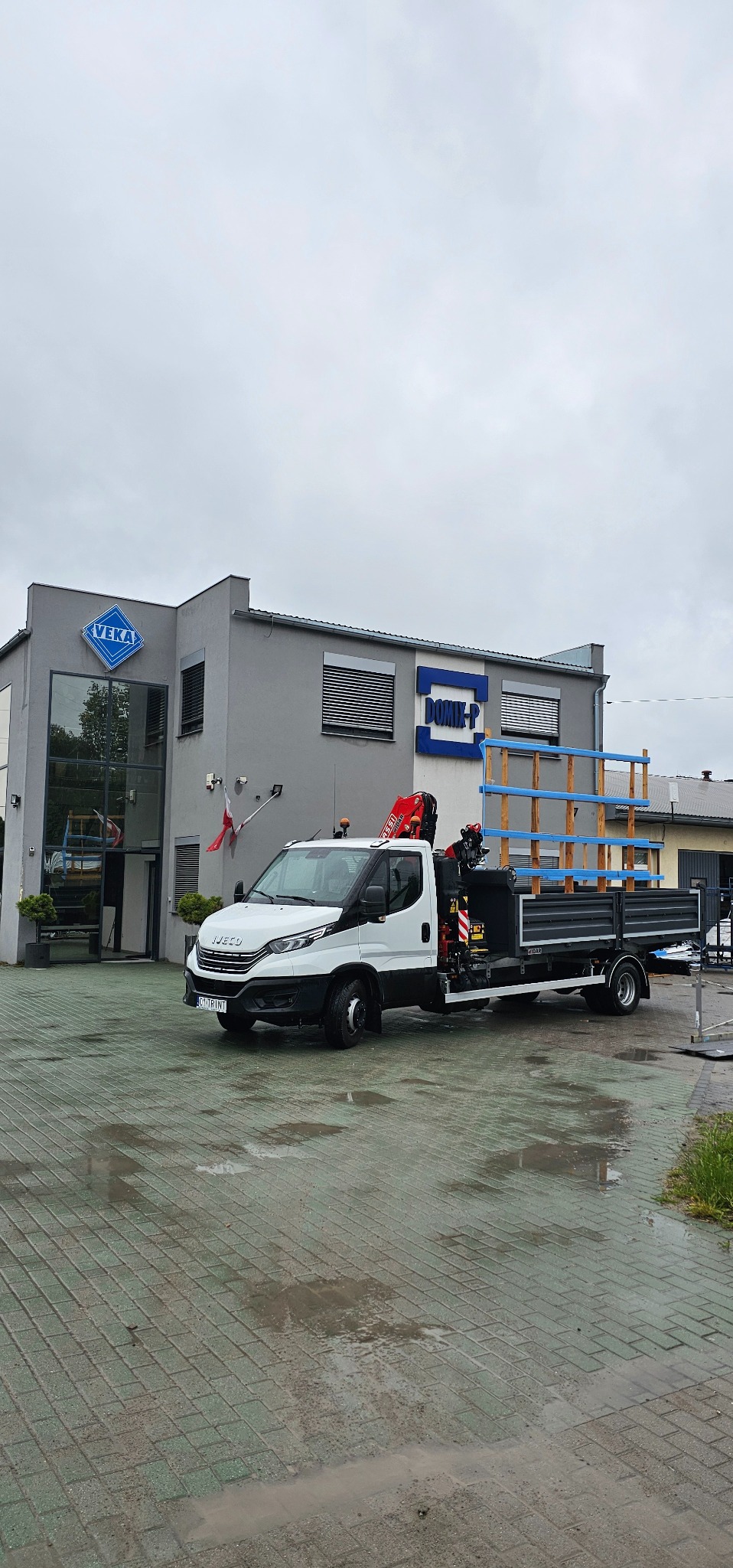 Biała ciężarówka z dźwigiem HDS na tle budynku firmy Veka i Domik-P. Na skrzyni ładunkowej konstrukcja z niebieskimi elementami. Mokry bruk na pierwszym planie.