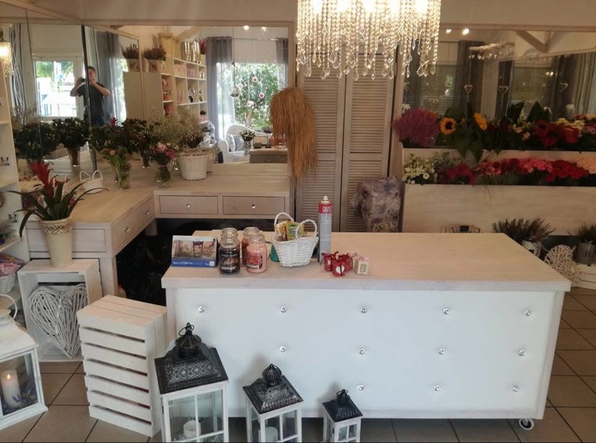 Jasne wnętrze kwiaciarni z drewnianymi meblami, kwiatami i dekoracjami. Na pierwszym planie biała lada zdobiona kryształkami, a w tle lustro odbijające przestrzeń.