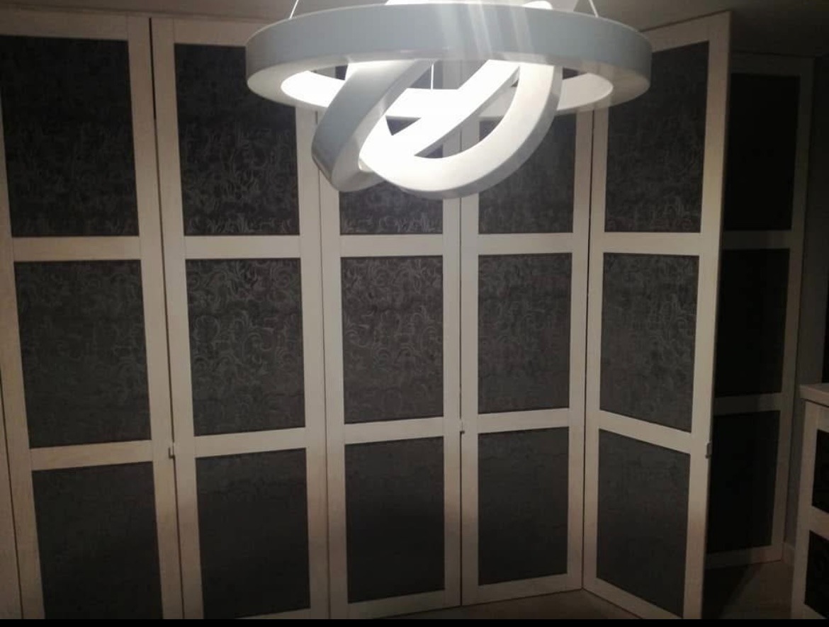 Białe, składane drzwi szafy z ciemnoszarymi, wzorzystymi panelami, oświetlone nowoczesną lampą. Jedne drzwi są lekko otwarte, ukazując wnętrze szafy.