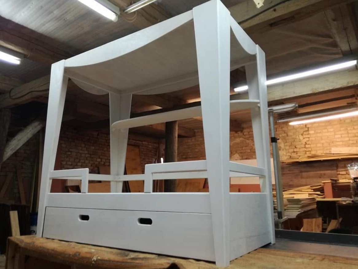 Białe, drewniane łóżko z baldachimem i szufladami, ustawione w warsztacie na drewnianym stole. Widoczne ceglane ściany i drewniana konstrukcja dachu.