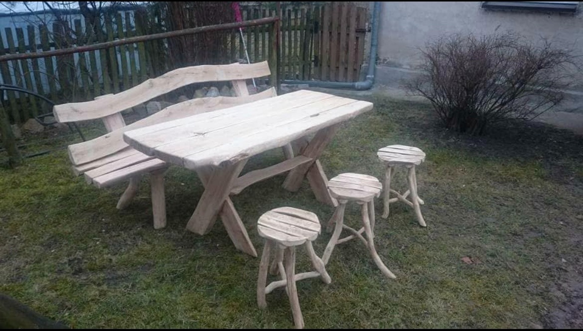 Rustykalny komplet ogrodowy: stół z ławką i stołkami z surowego drewna, ustawiony na trawie. Nierówne krawędzie blatu i naturalne kształty nóg podkreślają unikalny charakter.