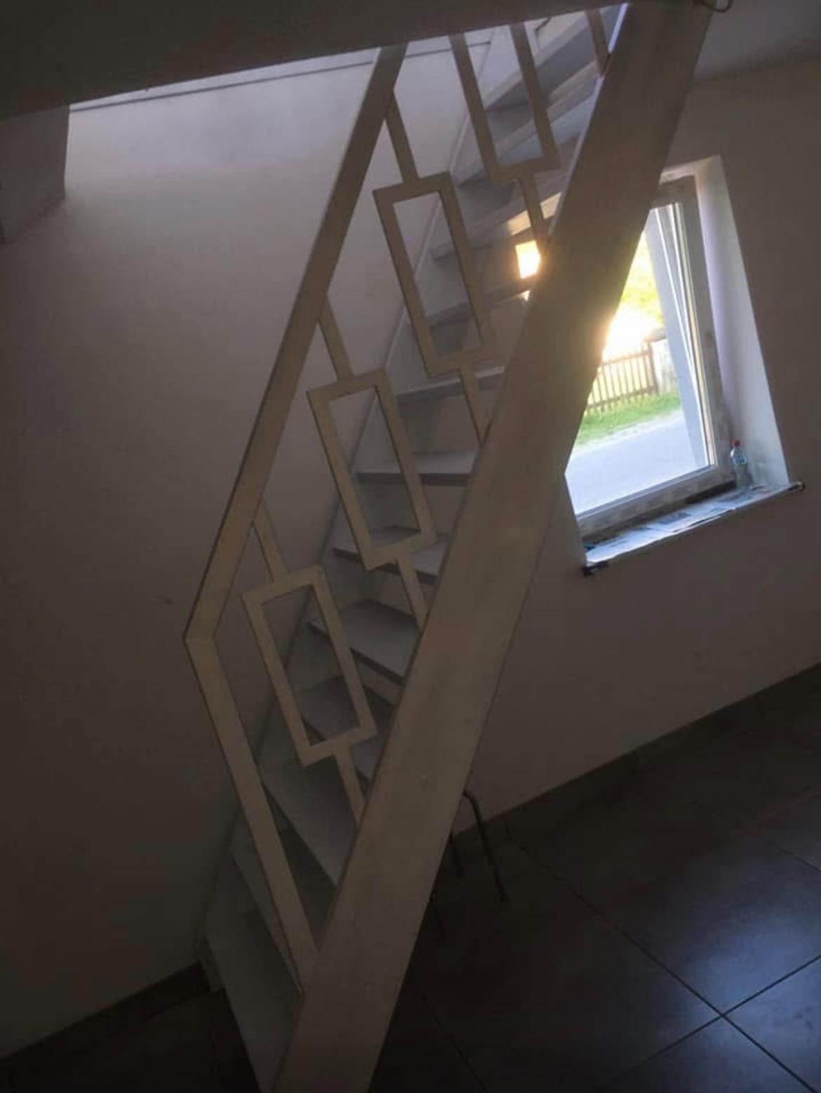 Białe schody z ozdobną balustradą z geometrycznym wzorem, widok z dołu. W tle okno z widokiem na zewnątrz, wpadające światło słoneczne. Minimalistyczny design.