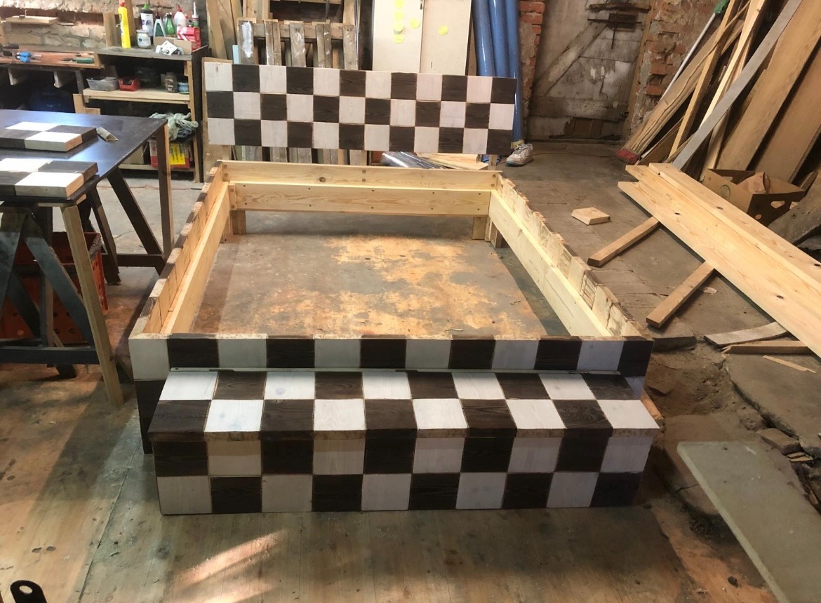 Niedokończona drewniana konstrukcja w szachownicę w warsztacie, z widocznymi deskami i narzędziami, sugerująca etap budowy lub renowacji mebla.