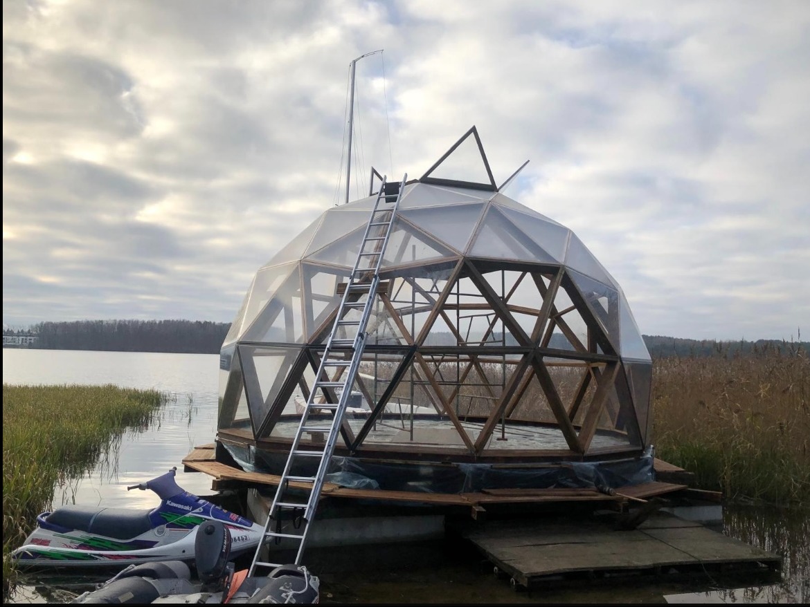 Geodezyjna kopuła na platformie pływającej na jeziorze, z drabinką i skuterem wodnym. Konstrukcja z drewna i przezroczystych paneli w pochmurny dzień.