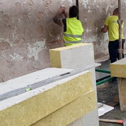 MIROSŁAW OCZKOWSKI KAMIR-BUD - Pracownicy w żółtych kamizelkach montują ocieplenie na elewacji budynku ze zniszczonym tynkiem. Widoczne rusztowanie, materiały izolacyjne i narzędzia.