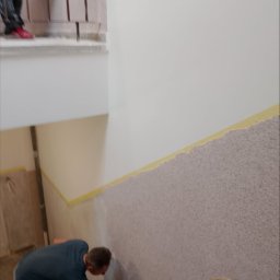 MIROSŁAW OCZKOWSKI KAMIR-BUD - Nakładanie dekoracyjnej struktury na ścianę klatki schodowej. Prace wykończeniowe z użyciem szpachelki, widoczne taśmy malarskie i częściowo wykończona powierzchnia.