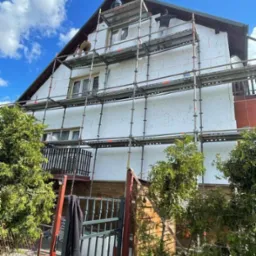 Dom w trakcie ocieplania, widoczne rusztowania i pracownicy przy pracy. Częściowo ocieplona elewacja styropianem, błękitne niebo w tle.