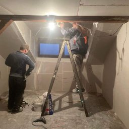 MIROSŁAW OCZKOWSKI KAMIR-BUD - Poddasze w trakcie wykończenia: pracownicy na drabinie montują płytę karton-gips, widoczne narzędzia i wiadro z gipsem. Surowy stan ścian, drewniane belki stropowe.