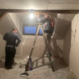 Poddasze w trakcie wykończenia: pracownicy na drabinie montują płytę karton-gips, widoczne narzędzia i wiadro z gipsem. Surowy stan ścian, drewniane belki stropowe.