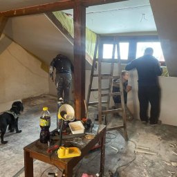 MIROSŁAW OCZKOWSKI KAMIR-BUD - Montaż płyt karton-gips na poddaszu z drewnianą konstrukcją. Widoczny pies, drabina i narzędzia pracy w pomieszczeniu ze skosami.