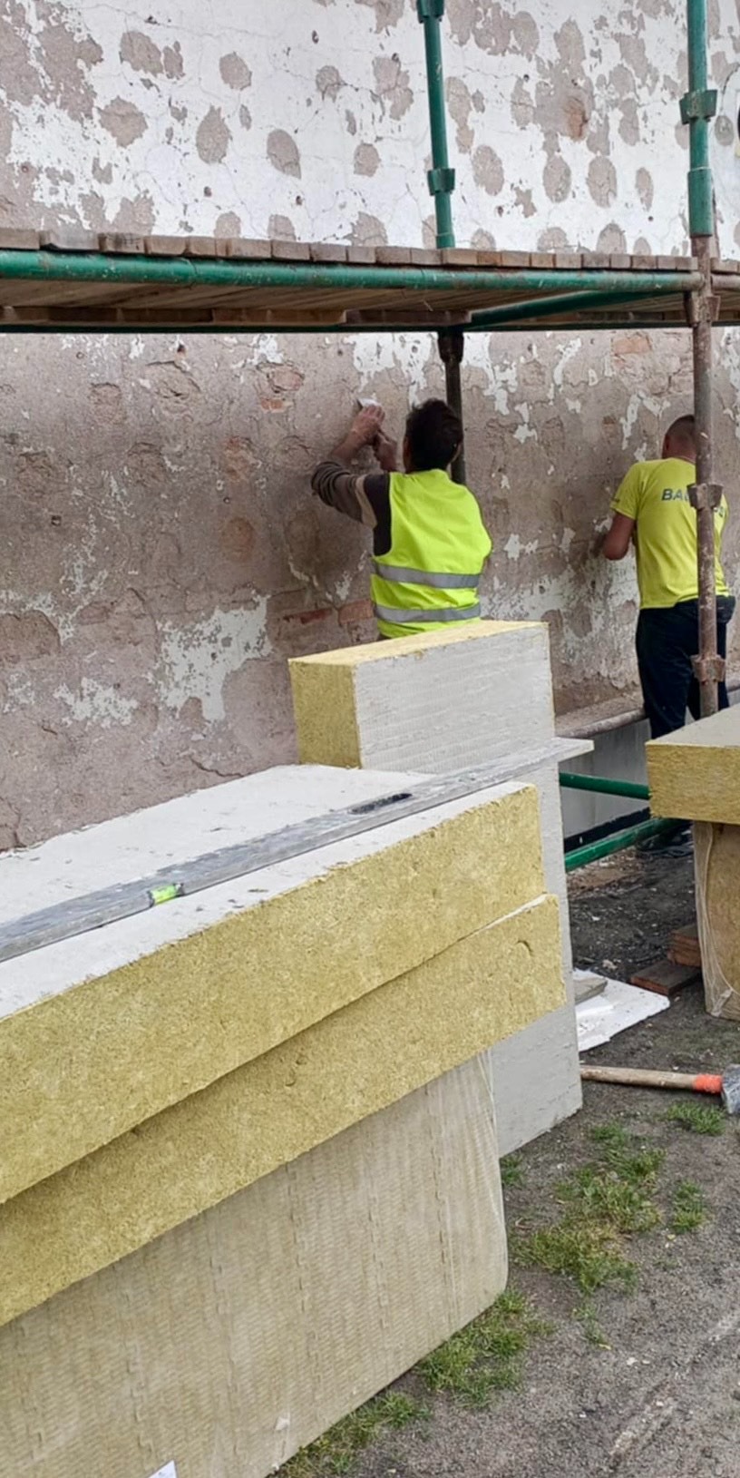 Pracownicy w żółtych kamizelkach montują ocieplenie na elewacji budynku ze zniszczonym tynkiem. Widoczne rusztowanie, materiały izolacyjne i narzędzia.