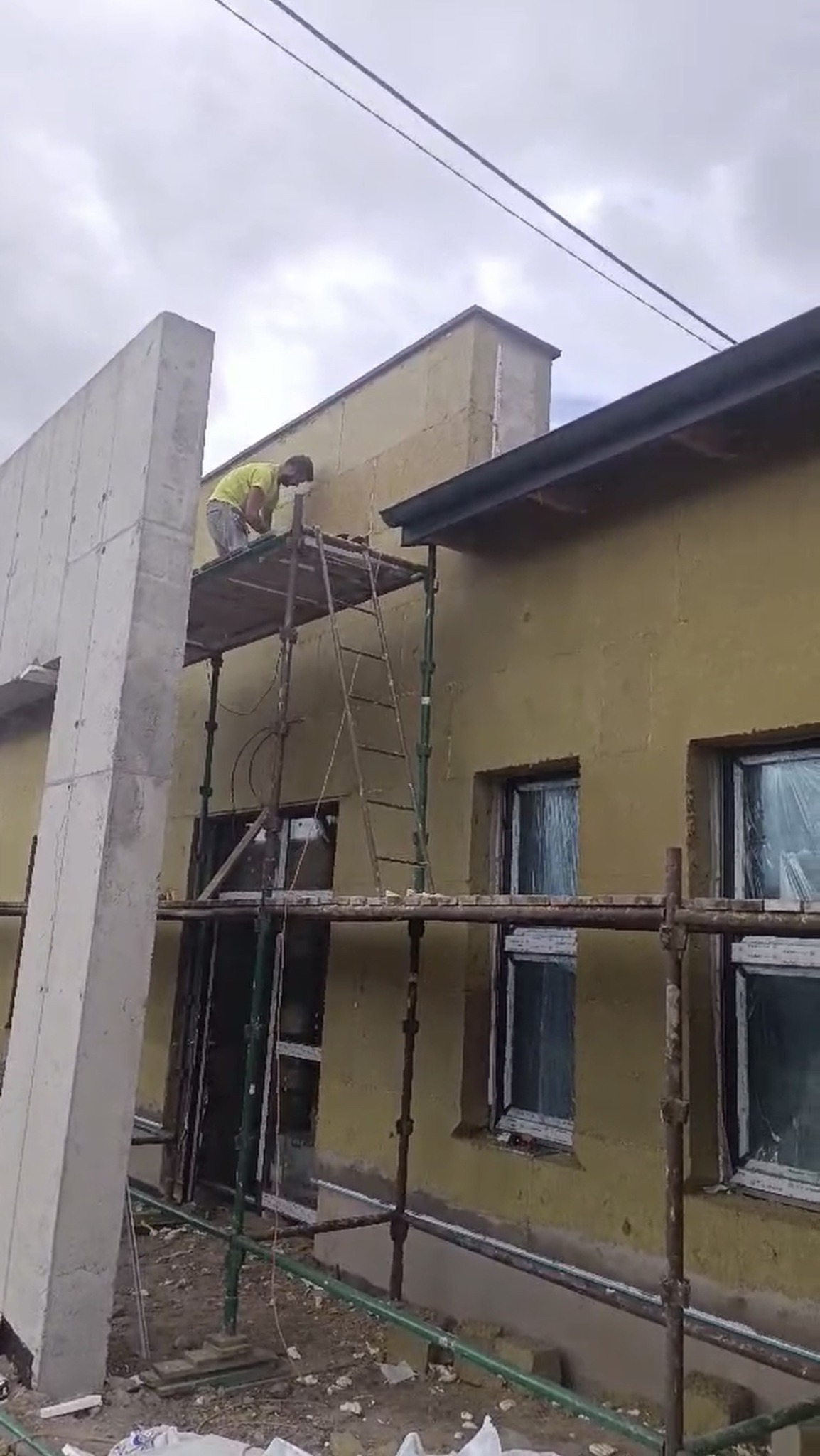 Pracownik na rusztowaniu, ociepla ścianę budynku z oknami. Widoczny fragment ściany z betonu. Budynek ocieplony, przygotowany do dalszych prac wykończeniowych.