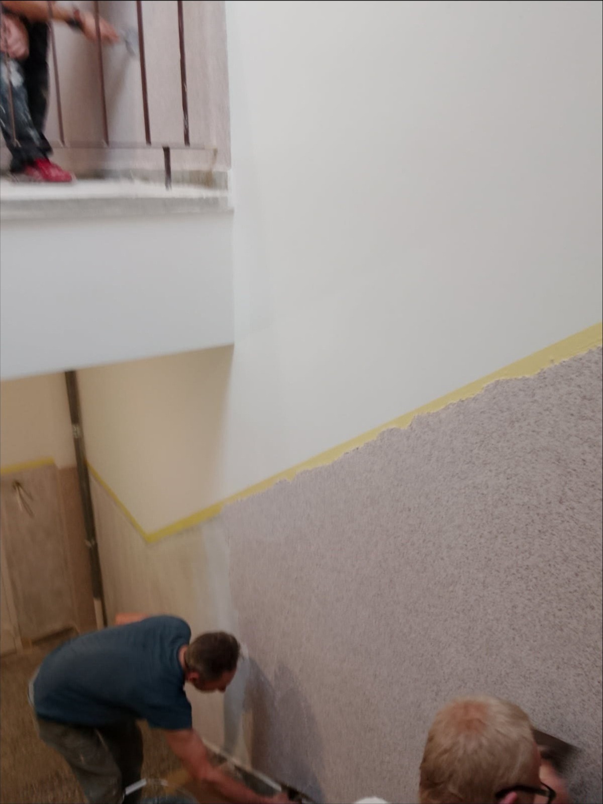 Nakładanie dekoracyjnej struktury na ścianę klatki schodowej. Prace wykończeniowe z użyciem szpachelki, widoczne taśmy malarskie i częściowo wykończona powierzchnia.