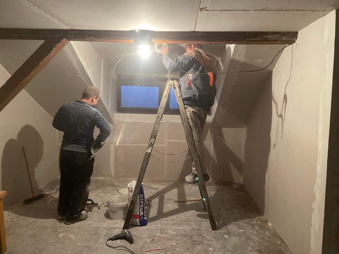 Poddasze w trakcie wykończenia: pracownicy na drabinie montują płytę karton-gips, widoczne narzędzia i wiadro z gipsem. Surowy stan ścian, drewniane belki stropowe.