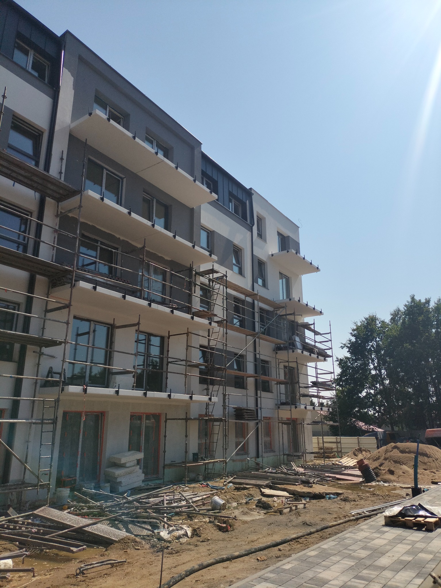 Budynek wielorodzinny w trakcie prac elewacyjnych, z widocznymi rusztowaniami i przygotowanym terenem. Jasne, słoneczne oświetlenie.