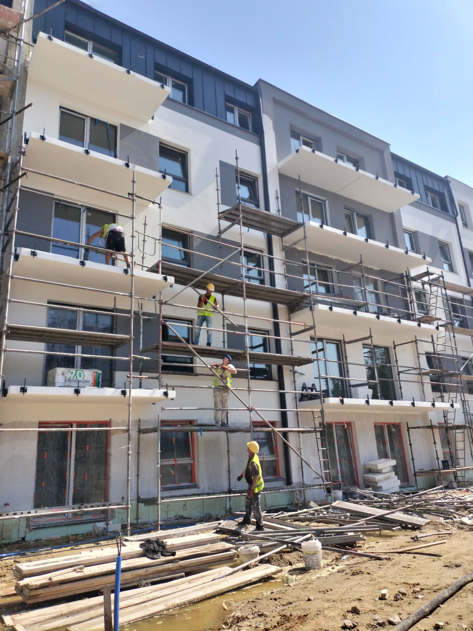 Prace elewacyjne na rusztowaniu przy budynku wielorodzinnym. Trzech pracowników w żółtych kaskach. Częściowo ocieplona elewacja w odcieniach szarości i bieli.