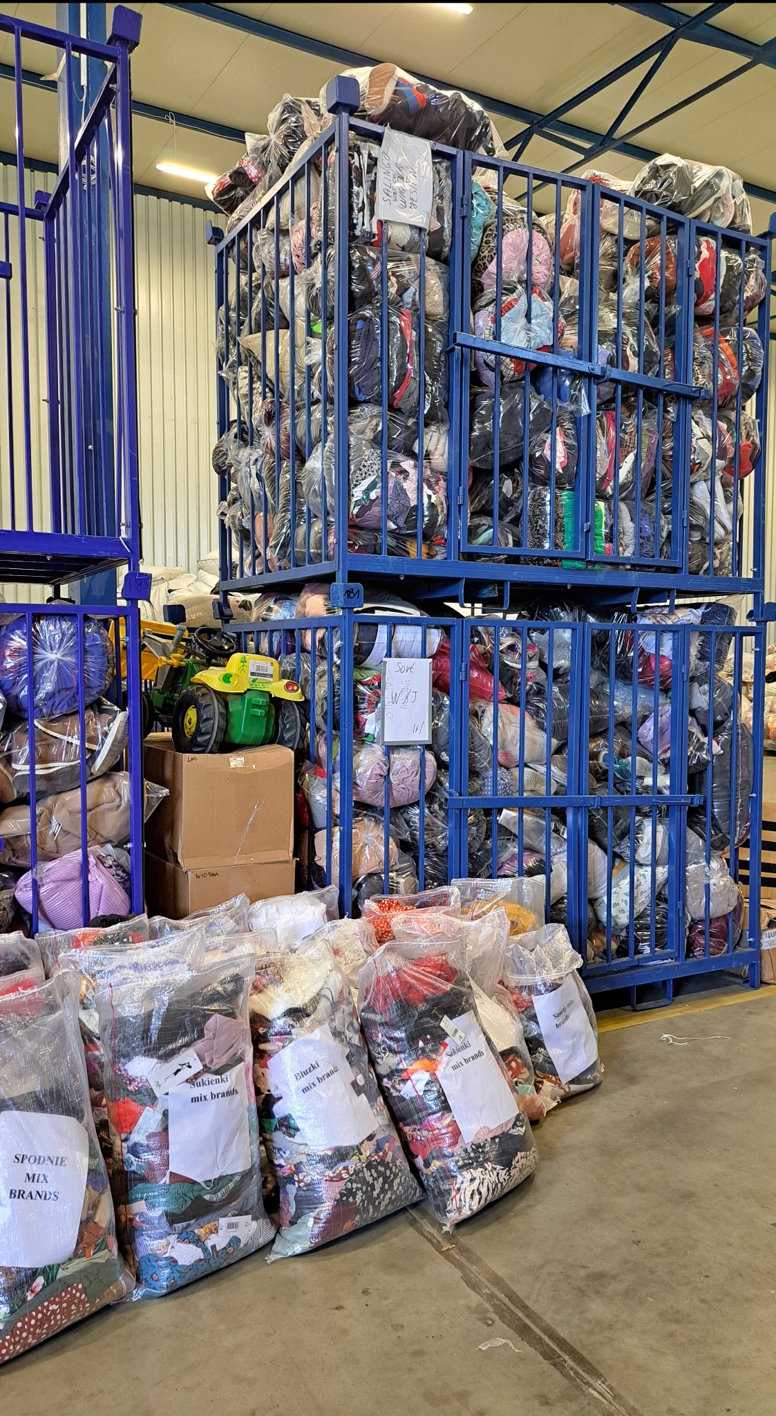 Magazyn odzieży używanej: metalowe kosze wypełnione workami z ubraniami, z etykietami 'spodnie mix brands', 'bluzki mix brand', 'sukienki mix brands'.