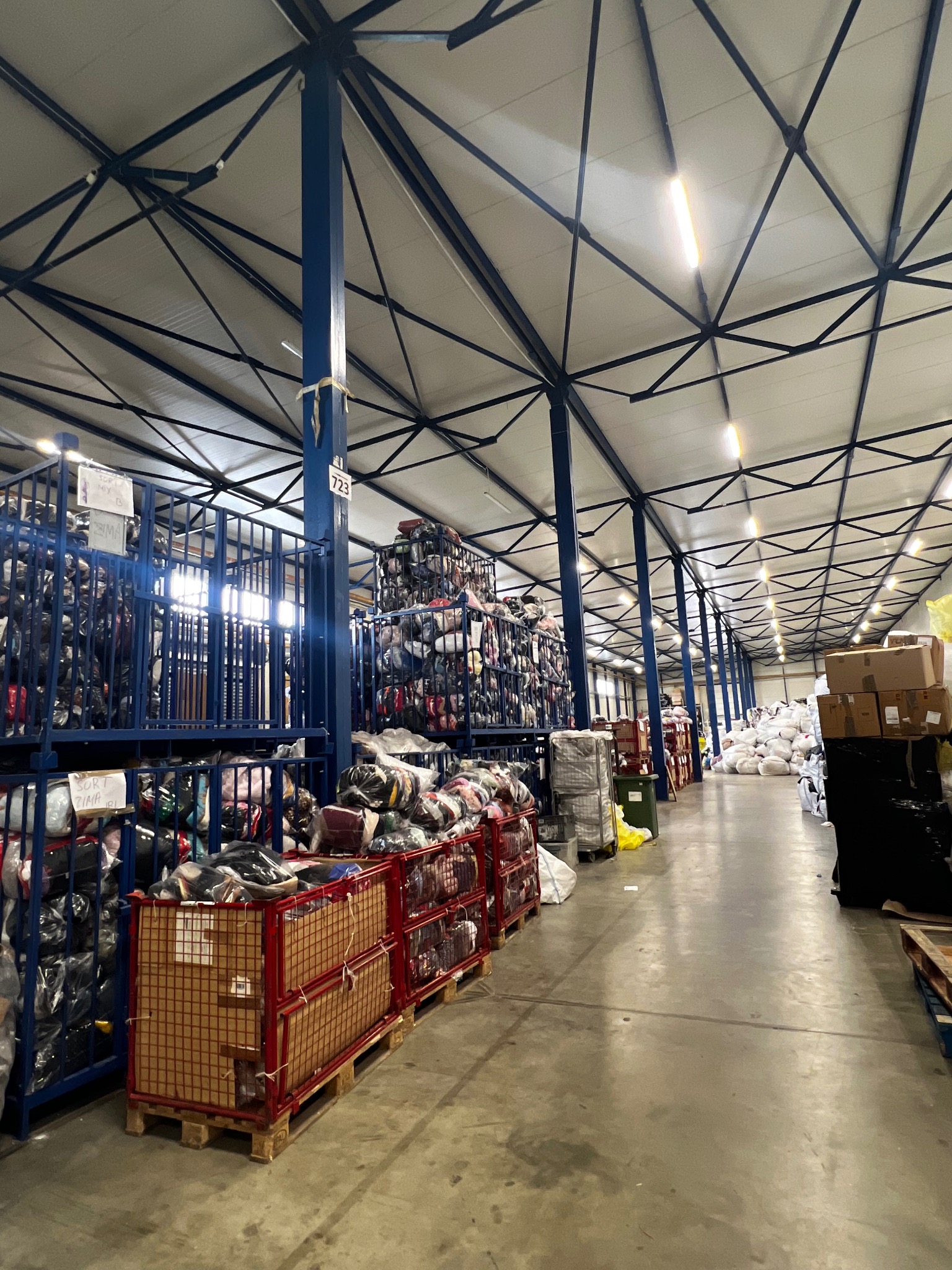 Magazyn odzieży używanej: regały wypełnione sortowaną odzieżą w metalowych koszach, oświetlone lampami sufitowymi, w tle sterty ubrań i kartonów.