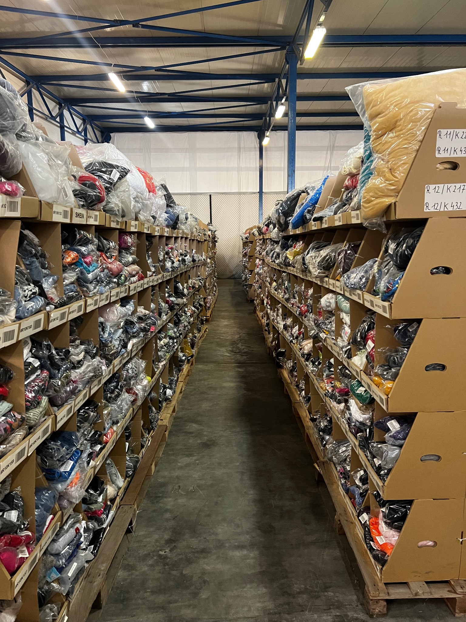 Magazyn odzieżowy: rzędy regałów wypełnione zapakowaną odzieżą, widok z perspektywy alejki. Etykiety na półkach. Oświetlenie magazynowe.