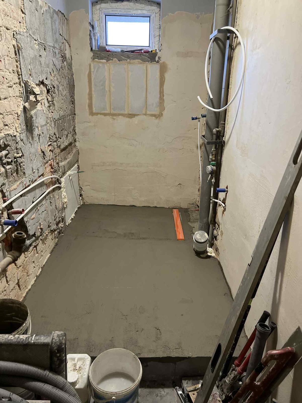 Wylewka samopoziomująca w łazience w trakcie remontu. Widoczne surowe ściany, instalacje i narzędzia. Pomieszczenie z oknem w stanie przygotowania do dalszych prac.