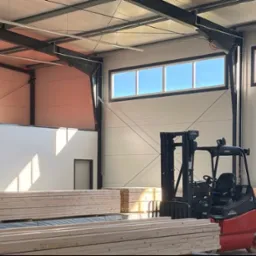 Wnętrze magazynu tarcicy: wózek widłowy Linde obok stosu jasnej tarcicy budowlanej pod oknami w hali z metalową konstrukcją.