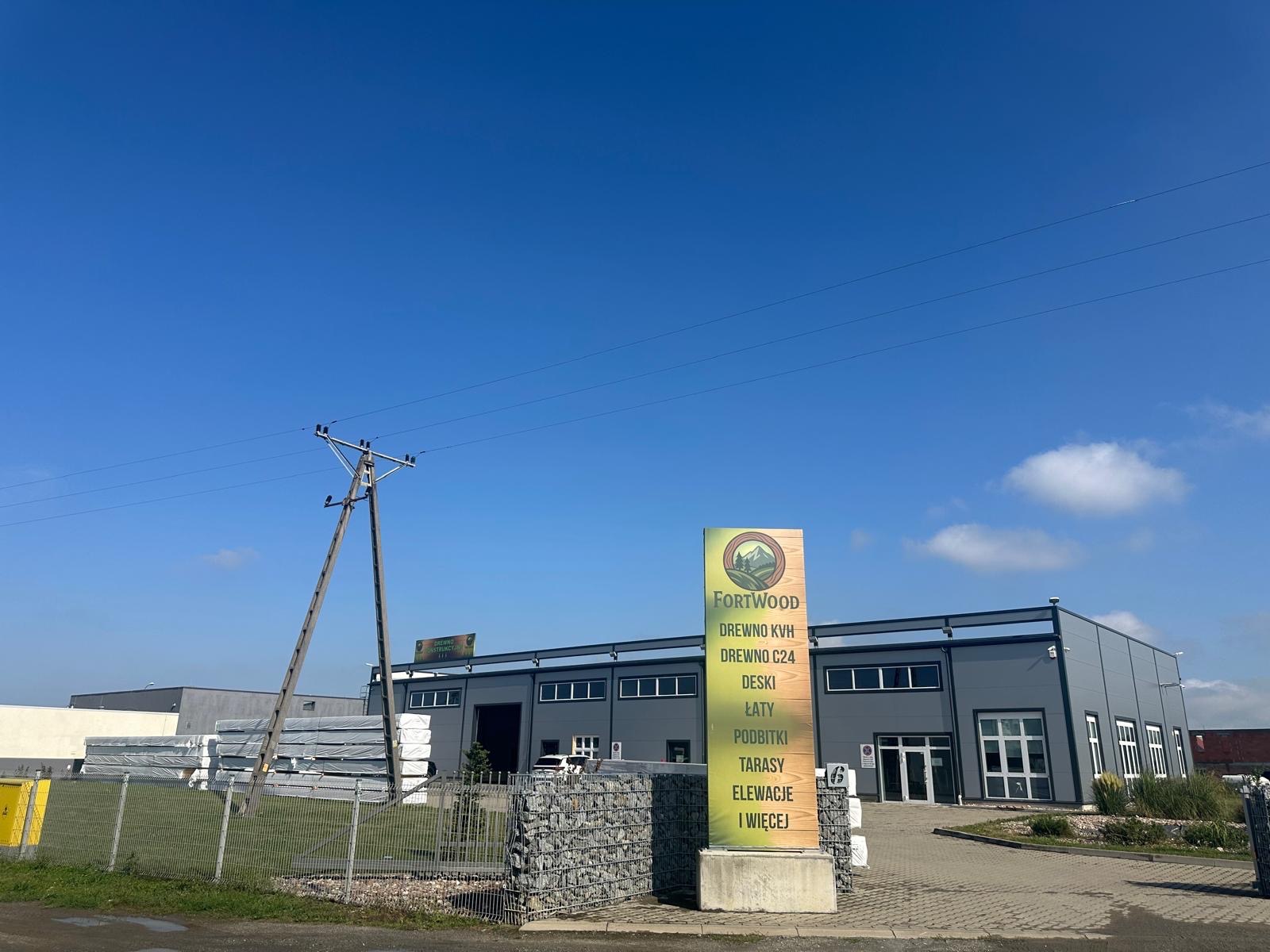 Siedziba firmy Fortwood z tablicą reklamową drewna KVH, C24, desek, łat, podbitki, tarasów i elewacji na tle szarego budynku magazynowego i błękitnego nieba.