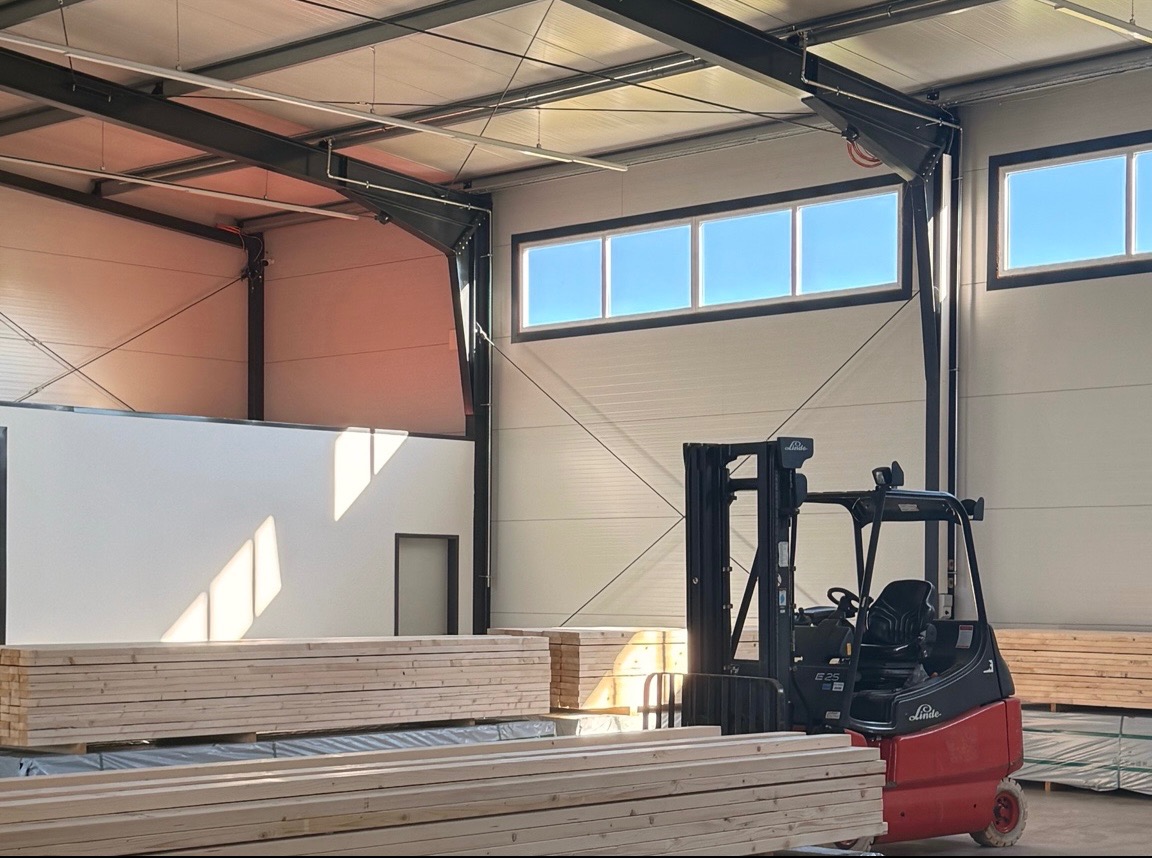 Wnętrze magazynu tarcicy: wózek widłowy Linde obok stosu jasnej tarcicy budowlanej pod oknami w hali z metalową konstrukcją.