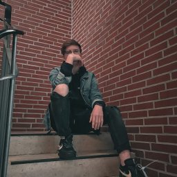 Albert Szymański - Młody mężczyzna w okularach, siedzący na schodach z ceglanym tłem, ubrany w jeansową kurtkę i czarne spodnie z dziurami na kolanach, pozujący do zdjęcia.