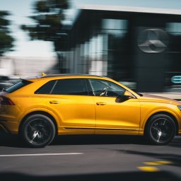 Albert Szymański - Dynamiczne ujęcie złotego Audi Q8 w ruchu, z rozmytym tłem miejskim i salonem Mercedesa w tle. Efektowna fotografia motoryzacyjna.