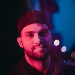 Portret mężczyzny w czapce z brodą, oświetlonego czerwonym i niebieskim światłem, z efektem bokeh w tle, tworzącym unikalną atmosferę. Nietypowe oświetlenie.