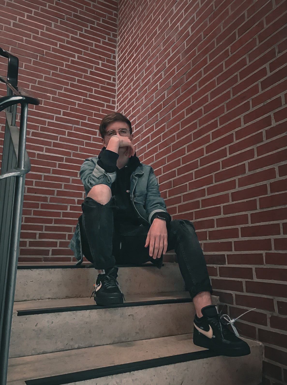 Młody mężczyzna w okularach, siedzący na schodach z ceglanym tłem, ubrany w jeansową kurtkę i czarne spodnie z dziurami na kolanach, pozujący do zdjęcia.