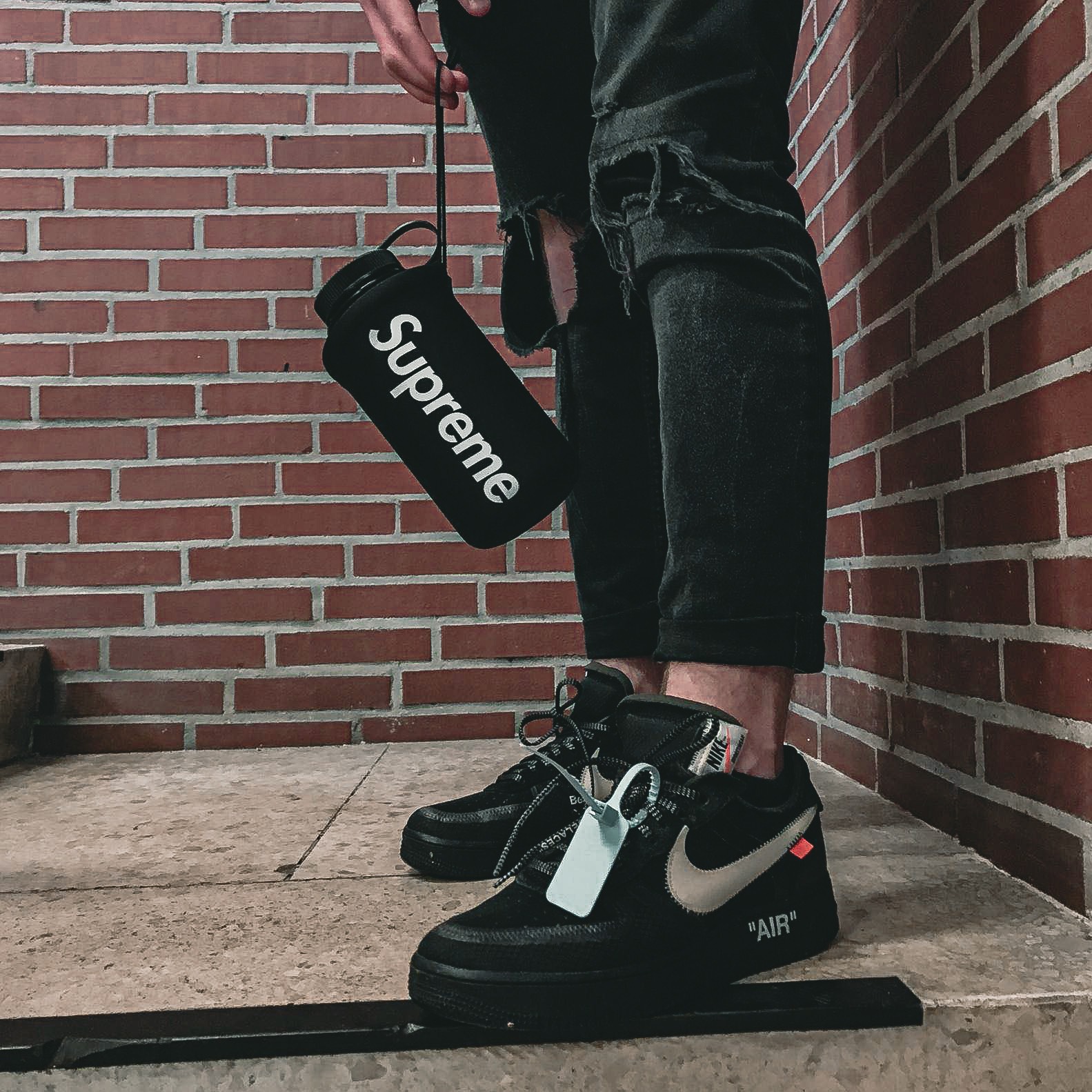 Osoba w czarnych, podartych jeansach i butach Nike Air Force 1 Off-White, trzymająca bidon Supreme na tle ceglanej ściany. Modny outfit.