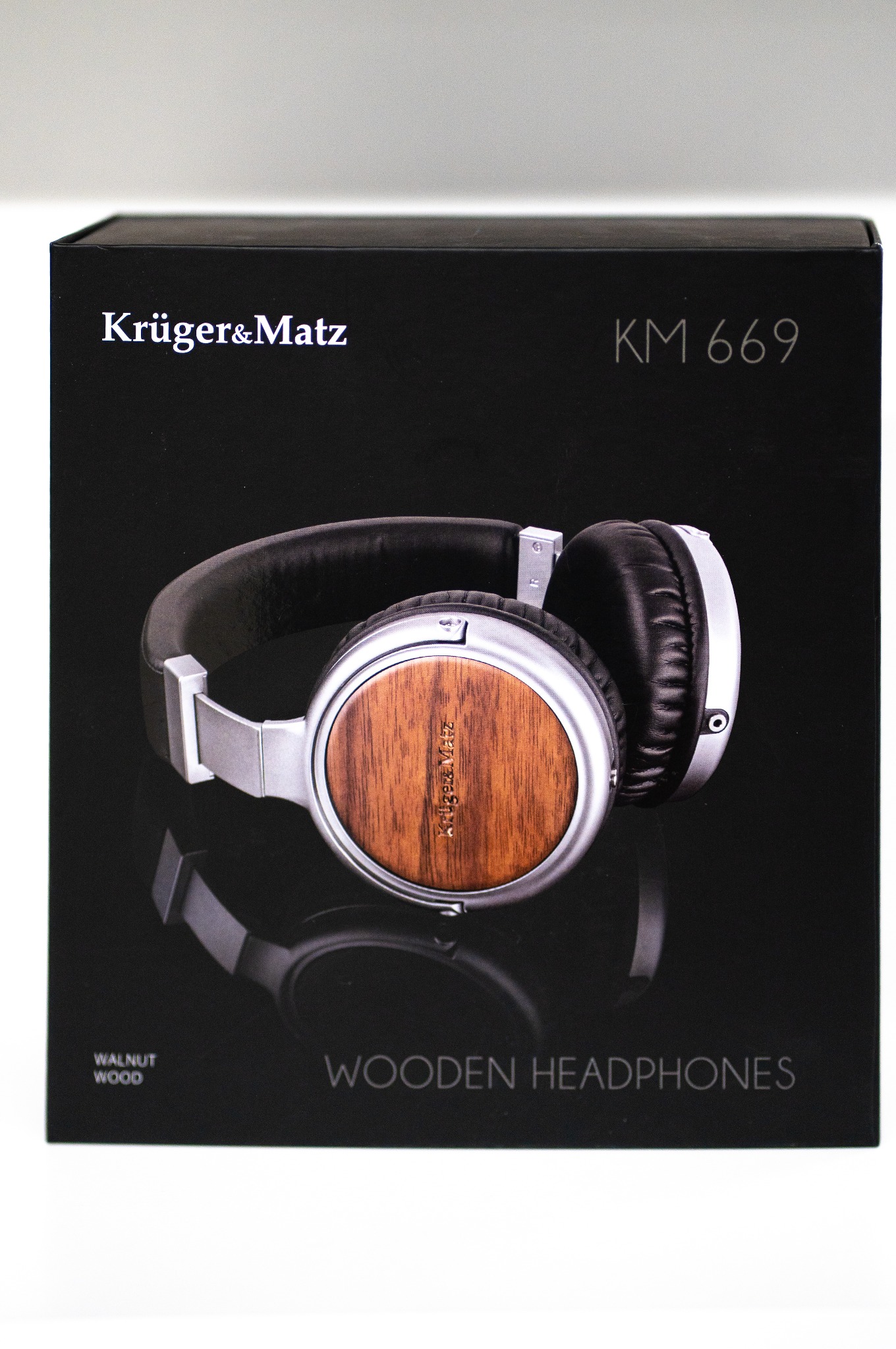 Eleganckie słuchawki Krüger&Matz KM 669 w pudełku. Model z drewnianymi nausznikami, srebrnymi elementami i czarnym, skórzanym wykończeniem pałąka. Widoczny napis 'Walnut Wood'.