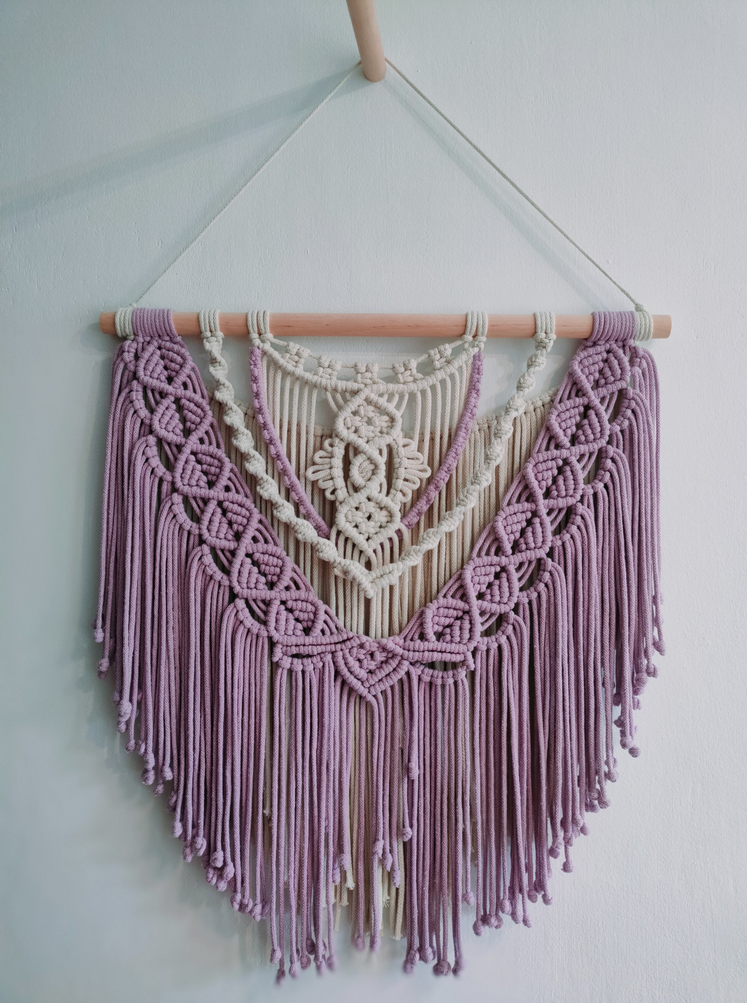 Ręcznie robiona makrama ścienna w odcieniach lila i beżu, zawieszona na drewnianym drążku, z dekoracyjnymi węzłami i frędzlami, na białym tle.