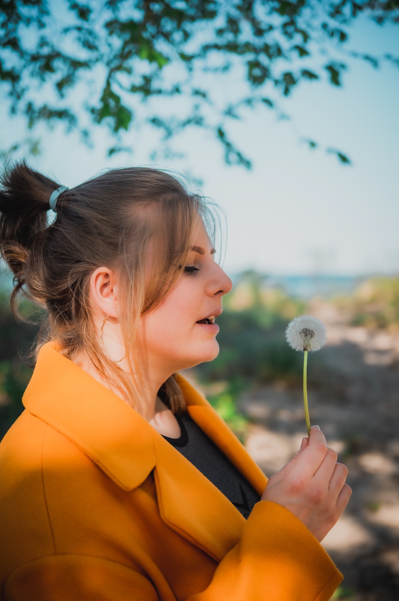 Profil kobiety w żółtym płaszczu trzymającej dmuchawca na tle rozmytego krajobrazu z drzewami i wodą, z delikatnym oświetleniem.