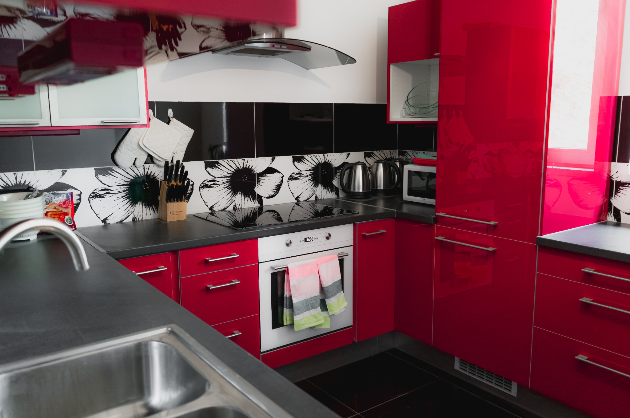Nowoczesna kuchnia w czerwieni z czarnym blatem i kwiatowym motywem na ścianie. Widoczny zlew, piekarnik, płyta grzewcza i okap. Stylizowane wnętrze.